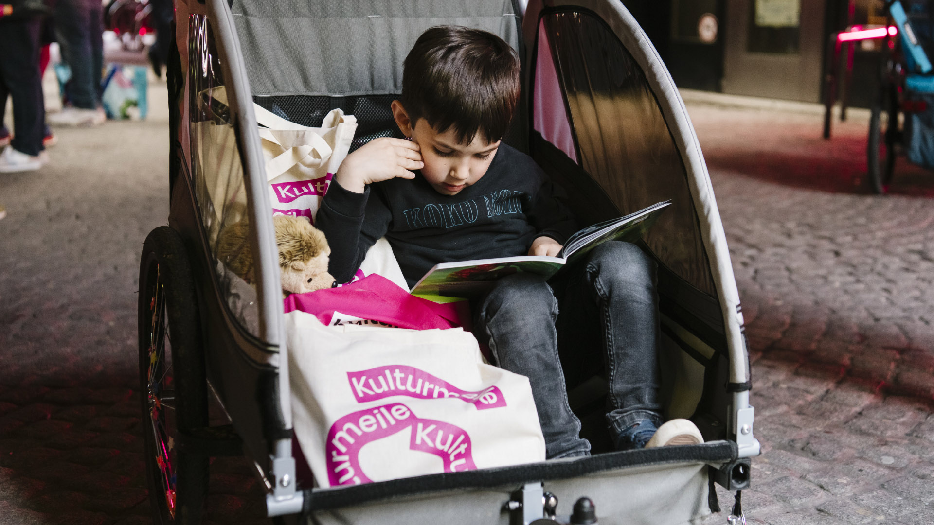 A little boy in a child trailer, next to him a couple of jute bags with Kulturmeile imprint