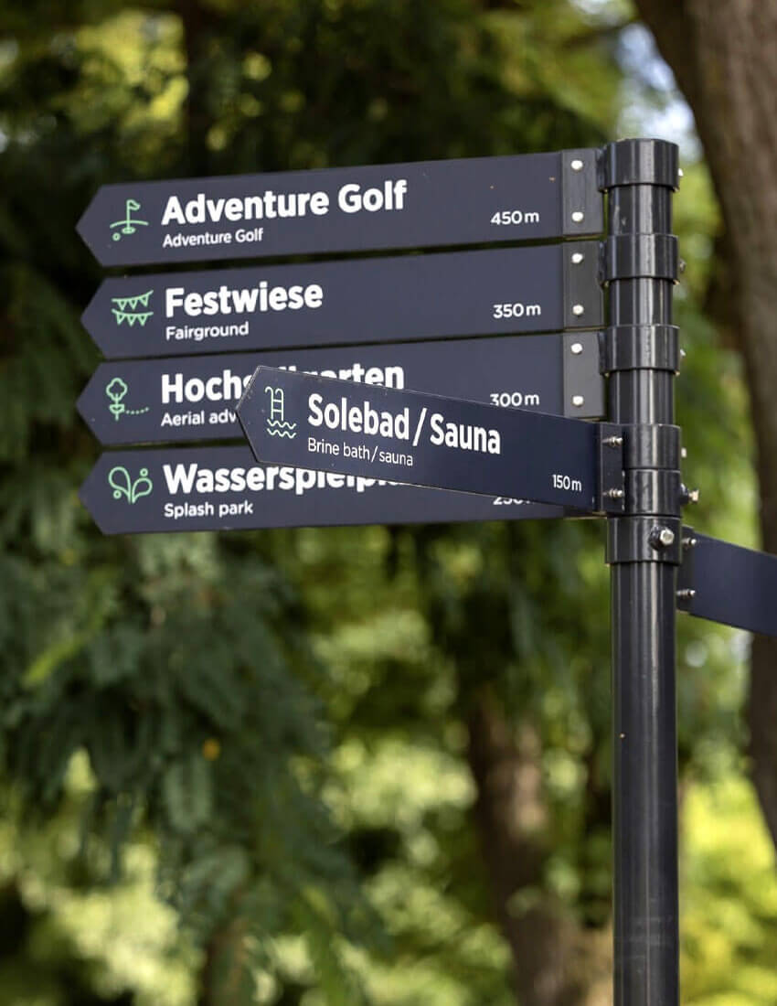 Bilingualer, moderner Wegweiser aus schwarzem Metall mit Schildern zu den Park-Attraktionen.