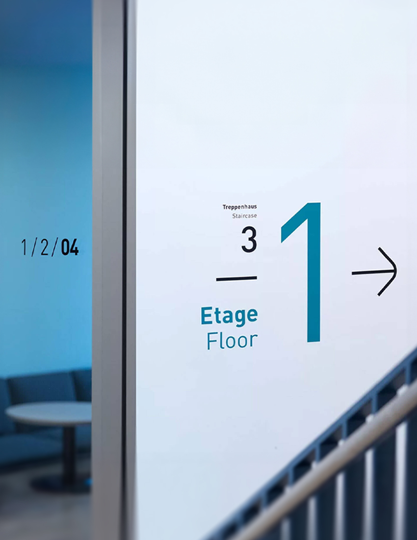 Beschilderung an der Wand mit großer Zahl „1“ und durchsichtiger Scheibe mit geplotteter Raumnummer, Blick auf dahinterliegendes Zimmer im Materna-Gebäude in Dortmund – Teil des Leitsystems von Florida Brand Design.